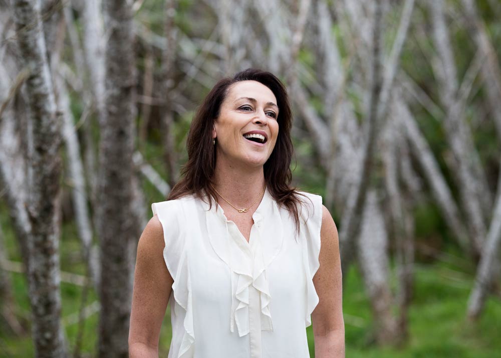 Susan standing amongst trees smiling in a white blouse