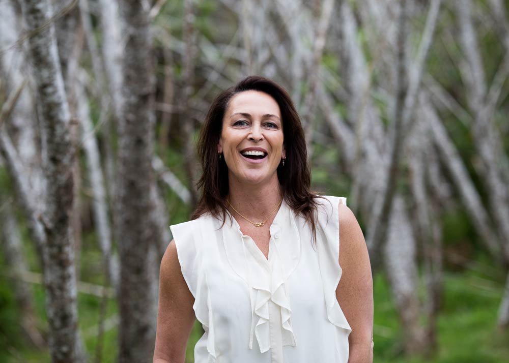 Susan standing amongst trees smiling in a white blouse