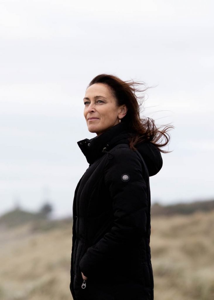 Susan looking out ahead on a beach, wearing a black jacket with the wind in her hair